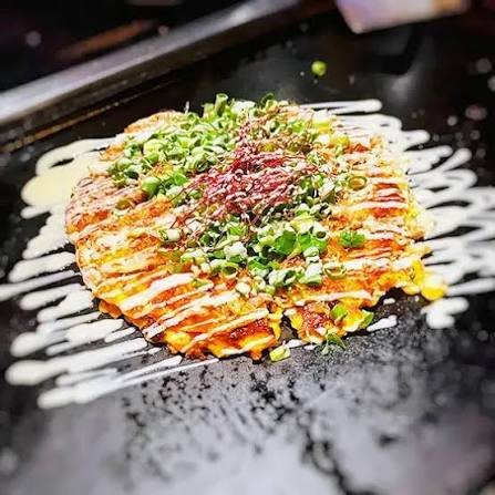 Een okonomiyaki op de bakplaat, met saus, mayonaise en lente-ui